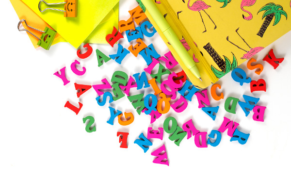 Várias letras coloridas, próximas a objetos escolares amarelos, sobre uma branca superfície plana.